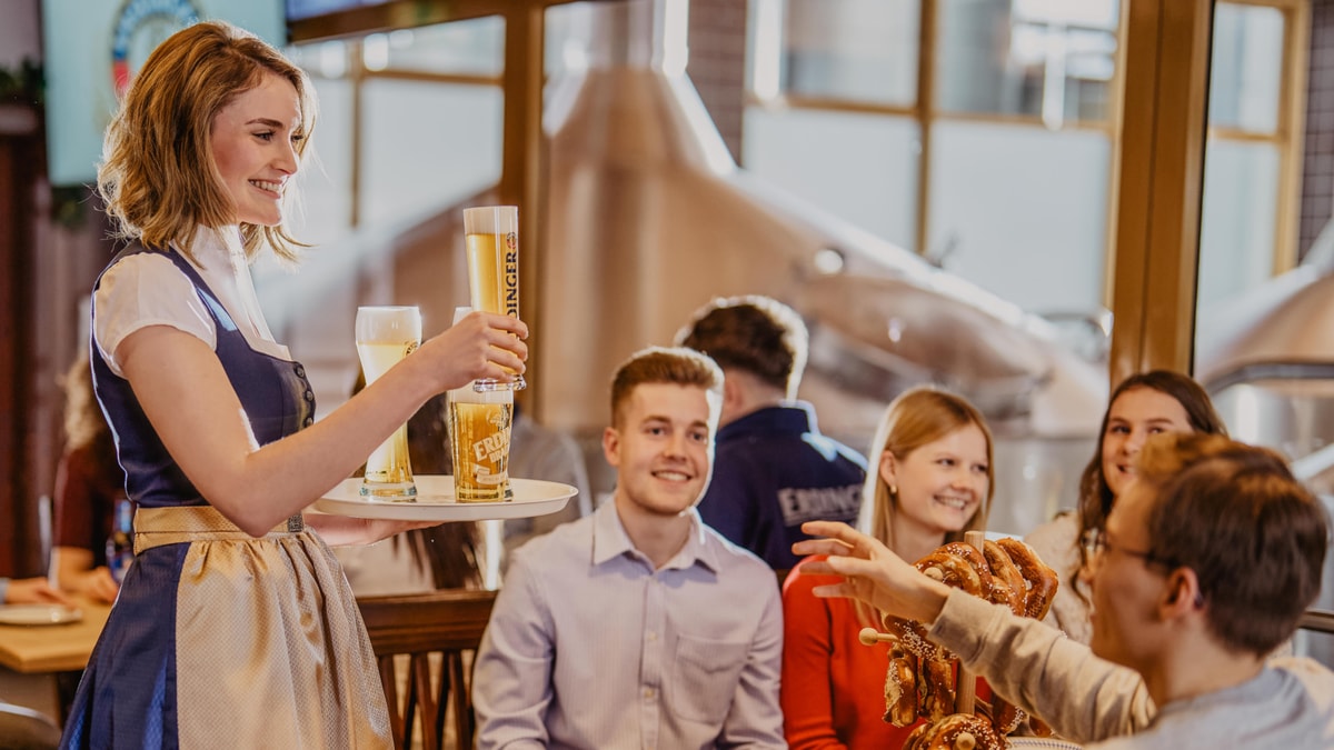 A waitress carries a tray with various ERDINGER beer specialties to smiling guests in the brewery.