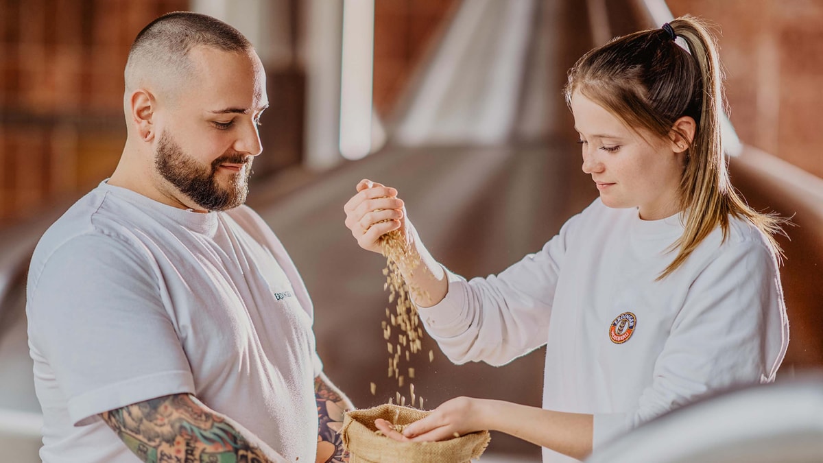 Two ERDINGER brewers with a sack of malt, the female brewer holds the malt grains in her hand and lets them fall into the sack.