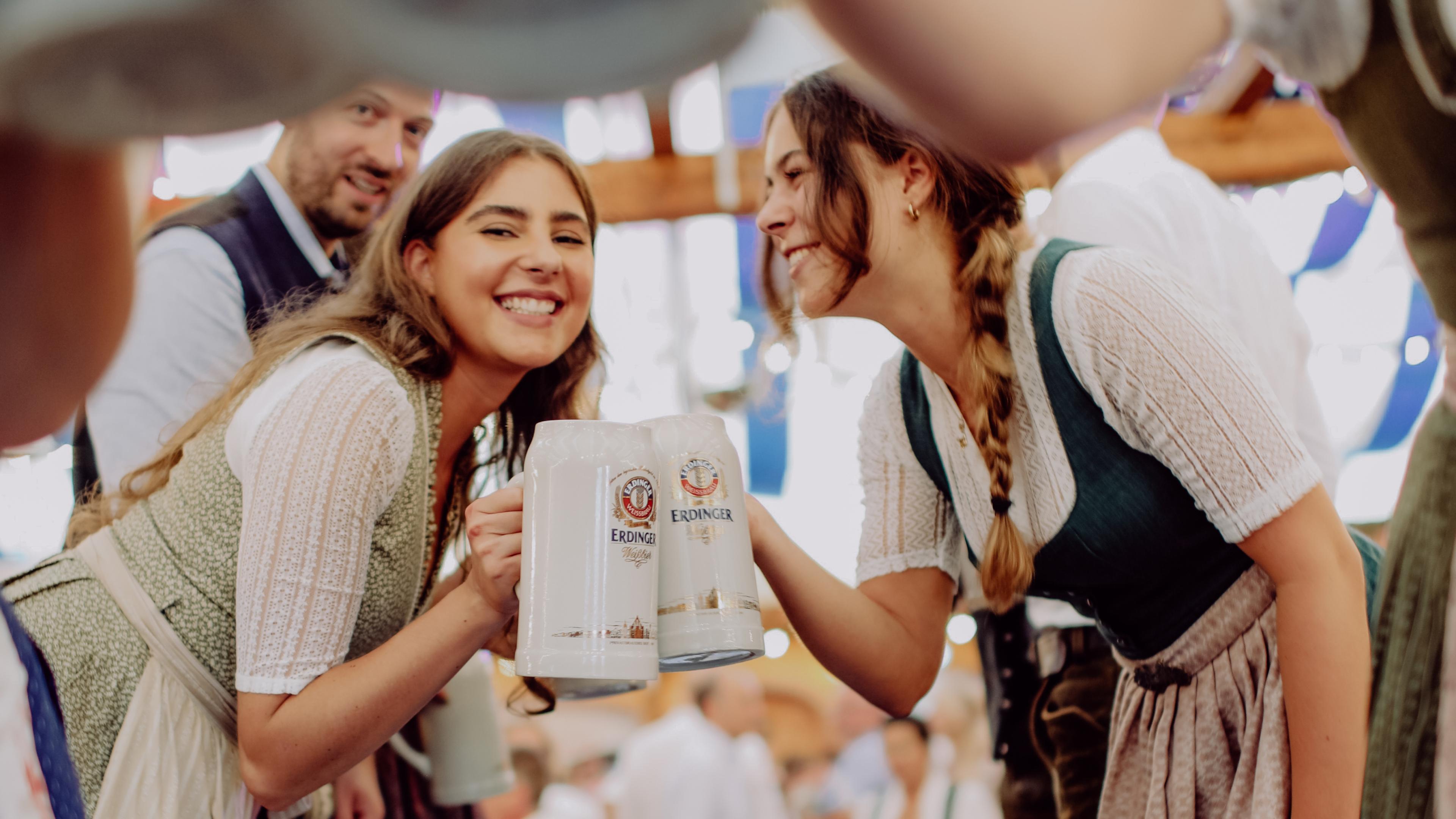 Zwei ERDINGER Mitarbeiterinnen in Tracht prosten sich mit Maßkrügen im Bierzelt zu auf dem Herbstfest der Brauerei.