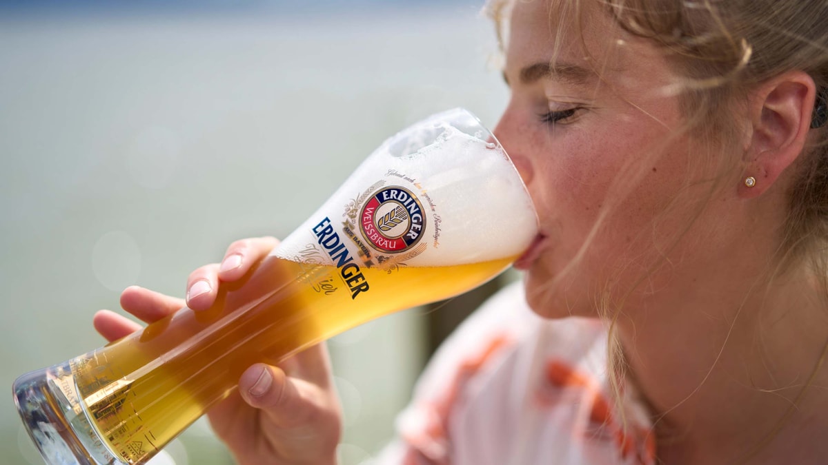 Eine Frau nimmt einen genüsslichen Schluck ERDINGER Weißbier an einem See.