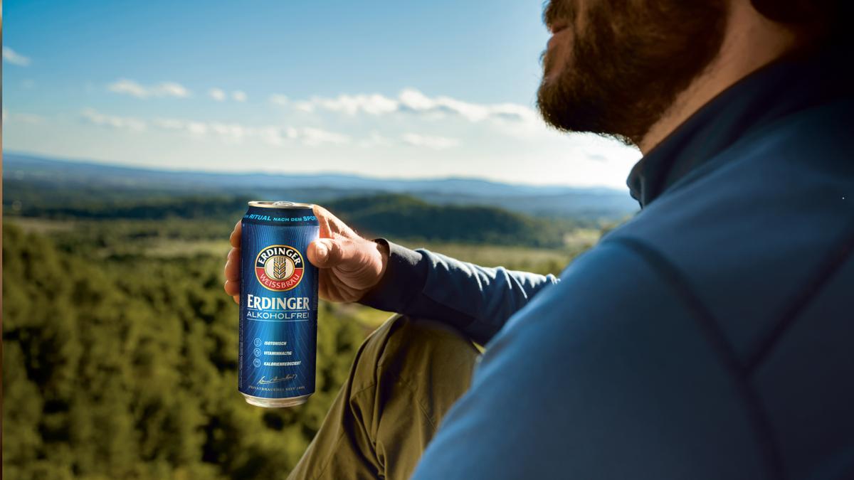 Ein Mann sitzt auf einem Gipfel und blickt in die Ferne mit einem ERDINGER Alkoholfrei in der Hand.