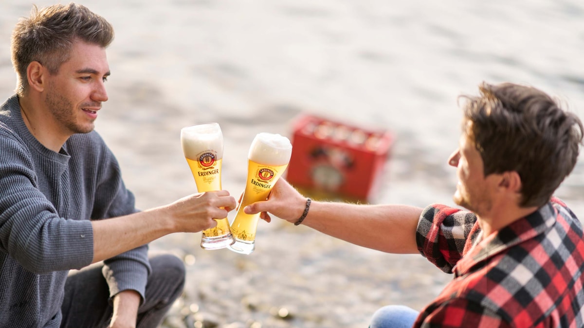 Zwei Männer prosten sich mit ERDINGER Weißbier an einem See zu.