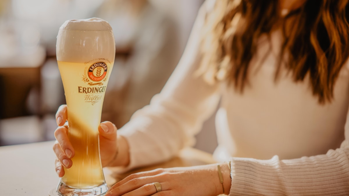Eine Frau greift ein ERDINGER Weißbier in einem Glas auf einem Tisch.
