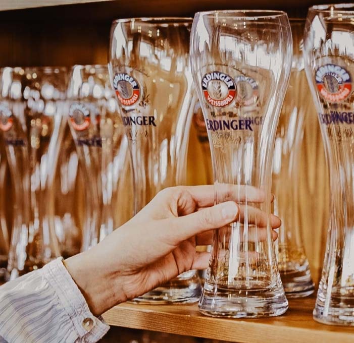 Eine Nahaufnahme von einer Hand die aus vielen aufgereihten ERDINGER Weißbiergläsern eines aus dem Regal nimmt.